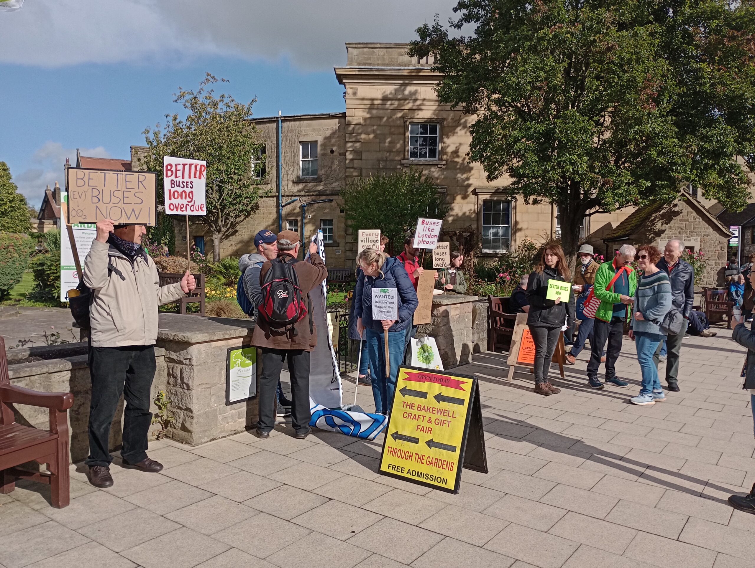 A group of people stand on the pavement holding placards demanding a better bus service